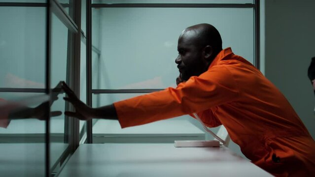 Side view of emotional Black prisoner sitting in visiting room, telling with beloved person on phone, holding hand on glass and saying goodbye, then guard taking him out