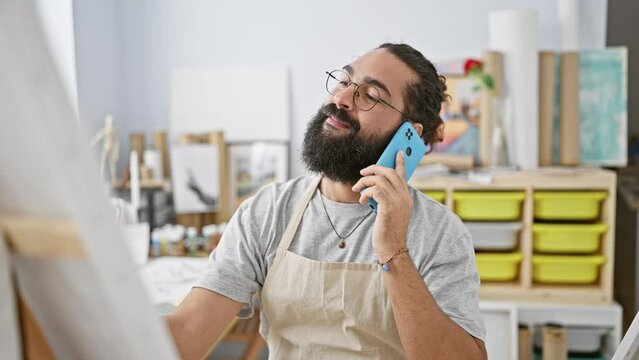 Smiling bearded man talking on blue smartphone in art studio with paintings and laptop