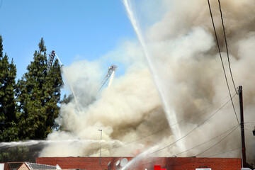 Firefighters battling a blazing building that is on fire with lots of smoke against blue sky background.