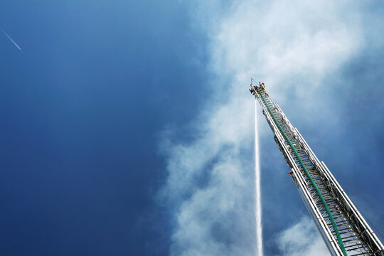 Horizontal image of an unmanned water canon on a hook and ladder fire engine shooting a stream of water onto a smoky structure fire with a blue sky background.