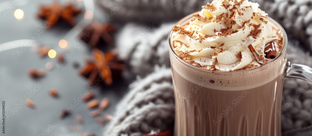 Wall mural Holiday drink with hot cocoa or coffee topped with whipped cream, shaved chocolate, spices, and a gray scarf, photographed against a blurred rustic background.