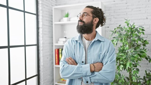 Confident bearded man with glasses standing in a modern office with folded arms.
