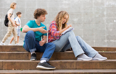 Teenage boy and girl sitting on steps near school and showing something by pointing index finger