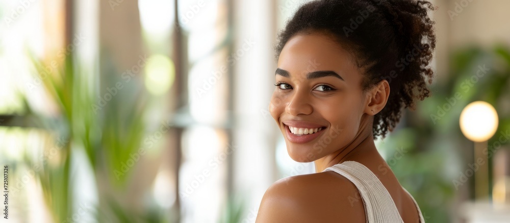 Wall mural Happy African American woman at home applying nourishing cream or lotion to her shoulder, with copy space.