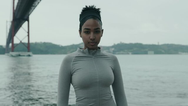 Sporty African woman stands by the river under a bridge, exuding motivation and confidence in activewear, set against a cloudy backdrop, ready for her run.