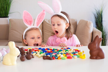 Small children, kids sisters with rabbit ears hunt for chocolate eggs eat them and laugh celebrate on the table in home.