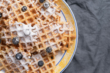 Waffles, blueberries, and powdered sugar on a yellow, blue, and white serving plate beside of a navy-blue linen napkin.