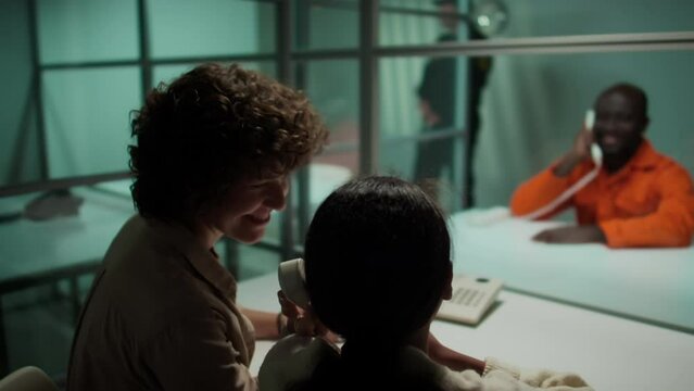 Wife and daughter telling supportive words on phone to arrested dad sitting behind glass wall in prison visiting room