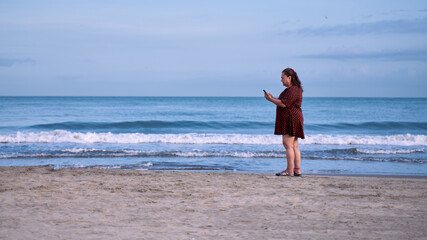 Fototapeta na wymiar Adult woman on the seashore standing on the beach looking at her phone on the beaches of Cartagena de Indias, Colombia.