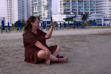 Woman in a beach dress sitting on the sand on the beaches of Cartagena taking a selfie. Tourist area of Cartagena de Indias, Colombia.
