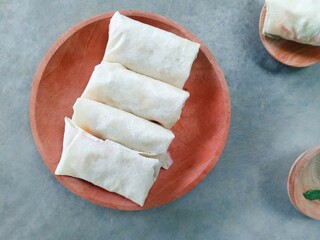 Lumpia, Spring rolls, traditional Indonesian snack filled with vegetables (cabbage, carrots and bean sprouts) served on a wooden plate on a gray background. fried food. uncooked spring rolls
