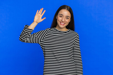 Young Caucasian woman smiling friendly at camera, waving hand gesturing hello greeting or goodbye welcoming with invitation hospitable expression. Pretty girl isolated on blue studio background