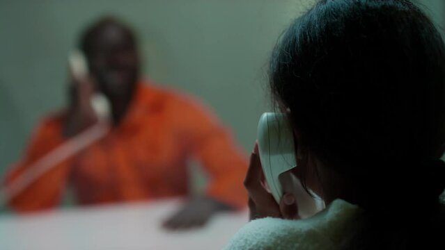 Foreground focus shot of girl sitting behind glass in visiting room and speaking with prisoner via landline phone