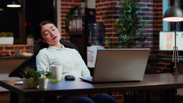 Sleepy employee asleep on computer desk at night in brick wall office, suddenly waking up. Tired businesswoman looking at watch, annoyed by time passing too slow while working overnight, camera B