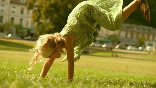 Little blonde girl performs handspring on a green park lawn on a sunny summer day