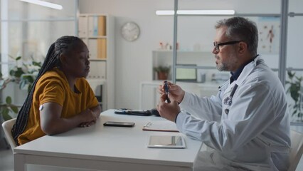 Medium shot of male doctor consulting new African American female patient with diabetes in clinic, demonstrating insulin pen for blood sugar control, teaching how to inject medication, woman listening