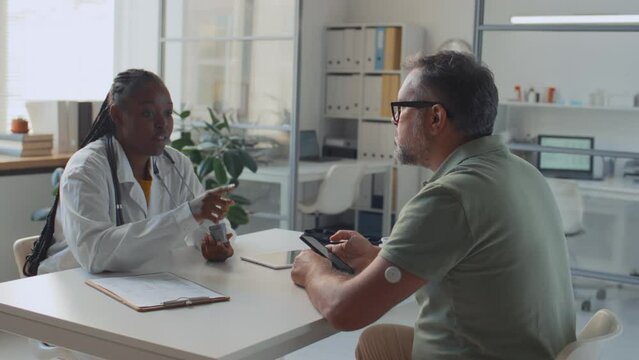 Medium shot of African American female GP teaching mature Caucasian man with diabetes how to use glucose monitoring device, implant sensor in arm, upload data to smartphone, while working in clinic