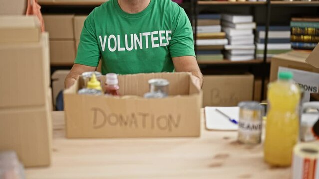 A young man in a green 'volunteer' t-shirt organizes food donations indoors at a charity event.