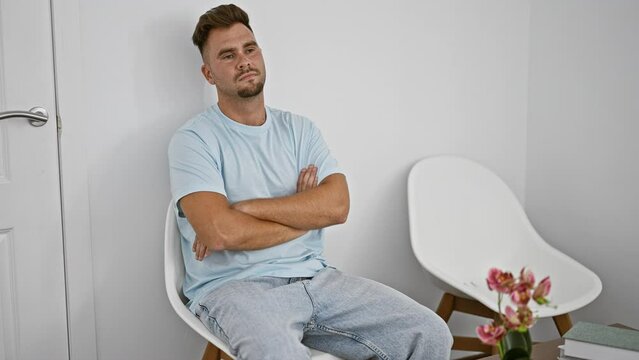 Serious young man with crossed arms sitting in a minimalistic white room with a modern chair and flowers.