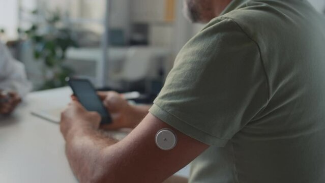 Medium close-up shot of middle-aged Caucasian man with diabetes sitting in GP clinic, after having glucose monitoring device implanted on arm, and learning to use blood sugar control app on smartphone