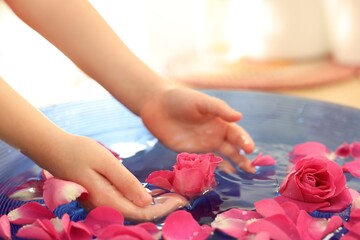 Child soaking hands in bowl with water and rose petals on blurred background, closeup