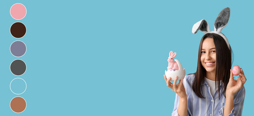 Pretty young woman with bunny ears, toy rabbit and Easter basket of eggs on light blue background....