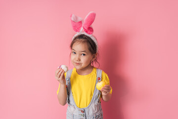Little girl with bunny ears on her head and Easter eggs in her hands against a pink background.