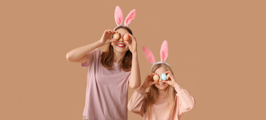 Funny mother and her little daughter with Easter eggs and bunny ears on beige background