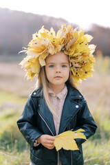 Little girl in a wreath of autumn leaves stands in a sunny meadow with a yellow leaf in her hands