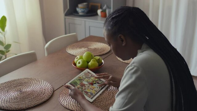 Medium high angle rear shot of young African American woman with long braids sitting at kitchen table at home, looking at picture with healthy foods on tablet computer while studying nutrition