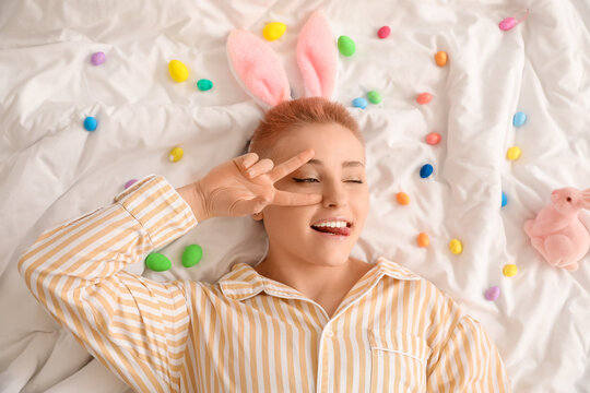 Young woman with Easter eggs and toy rabbits showing victory gesture on bed, top view