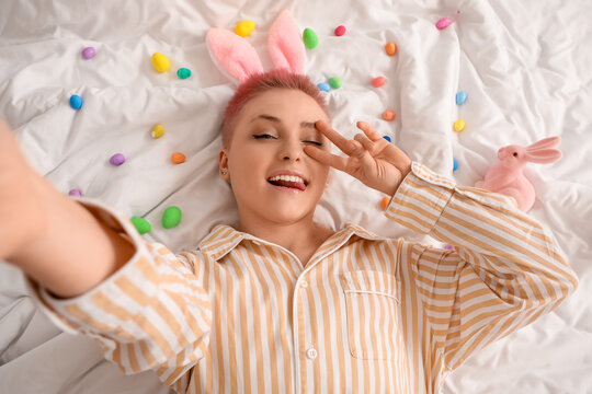 Young woman with Easter eggs and toy rabbits taking selfie on bed, top view