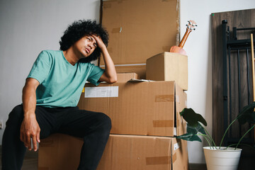 Asian man touching his head and sitting near stacked of carton boxes with sad expression