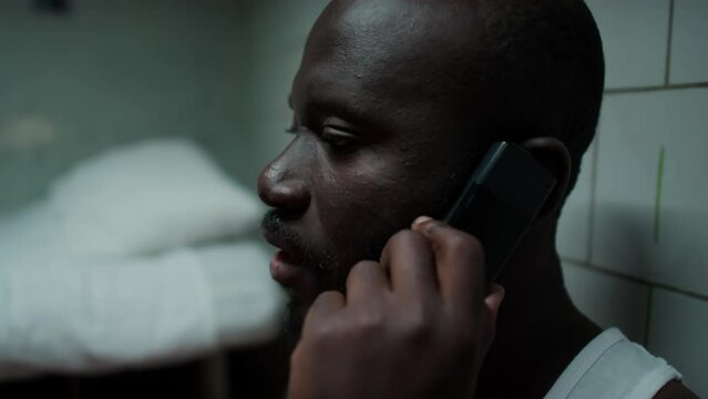 Tilt up shot of Black inmate sitting in prison cell, leaning on wall and having talk on mobile phone