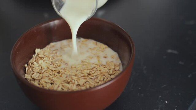 Close-up impersonal shot of mild being poured into brown breakfast bowl with dry oat flakes, on black kitchen counter