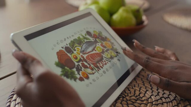 Hands of African American woman looking at picture on tablet computer on diabetic food, with fresh vegetables, fish, meat, avocado, broccoli, enlarging it and scrolling on touchscreen
