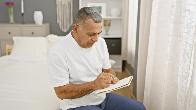 Mature hispanic man writing in notebook on bed in a well-appointed bedroom, evoking themes of planning or reflection.