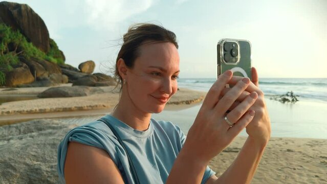 Pretty young caucasian woman takes pictures of the beautiful sea on tropical island beach. Action. Happy girl on vacation photographs on smartphone with palm trees on the background.