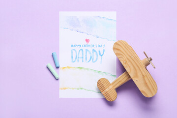 Card for Father's Day with chalks and wooden airplane on lilac background