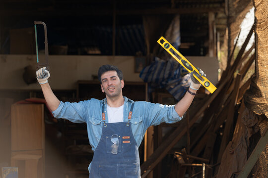 Portrait of a carpenter holding a spirit level and handsaw in his workshop.