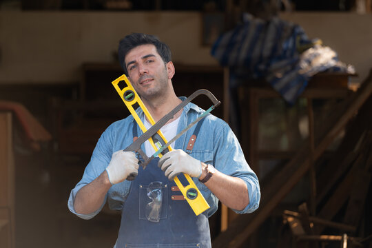 Portrait of a carpenter holding a spirit level and handsaw in his workshop.