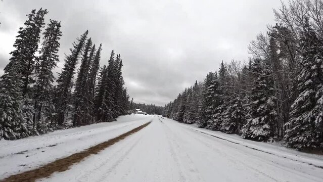 Hyperlapse POV from the front of a vehicle moving along a snow covered road through an evergreen forest. The sky is full of light gray clouds.
