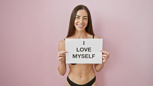 Beautiful, confident young hispanic woman in sexy lingerie stands looking proud over isolated pink background, holding a 'i love myself' banner - a stunning portrait of self-love and positivity