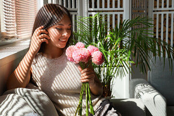 Beautiful young Asian woman with bouquet of pink dahlias sitting in living room near window