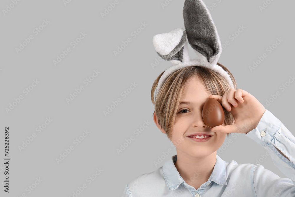 Wall mural cute little boy in bunny ears with chocolate egg on grey background. easter celebration