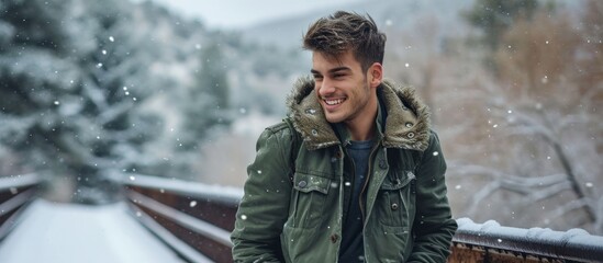 Stylish young man in a green bomber jacket enjoying snowy winter in Colorado on a bridge. - obrazy, fototapety, plakaty