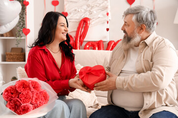 Beautiful mature couple with gift box and bouquet of roses sitting in decorated room. Valentine's...