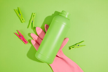 Colorful cleaning products on plain green background. A pink rubber glove holding a green bottle of detergent without label for design. Top view, house cleaning concept