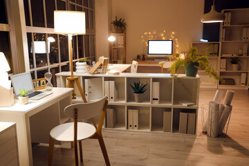 Interior of office with desks, shelf units and glowing lamps at night