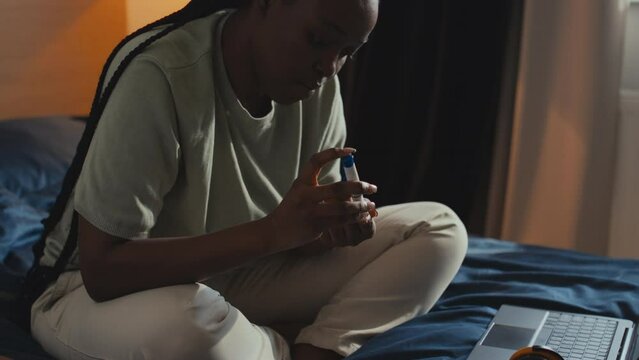 Medium shot of young black woman with diabetes sitting on bed, pricking finger with lancet, daubing blood onto test strip and inserting it into monitor, while playing ukulele at home